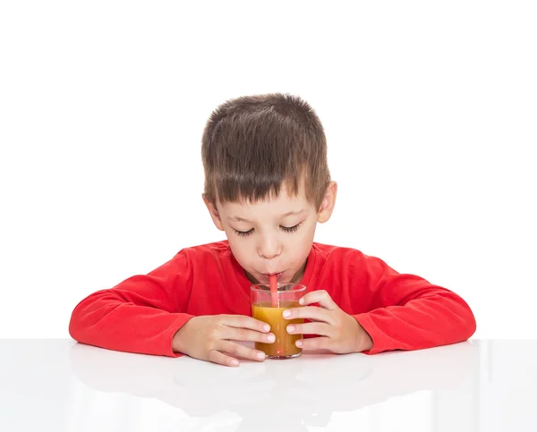 П'ятирічний хлопчик сидить за білим столом і п'є апельсиновий сік із соломи — стокове фото