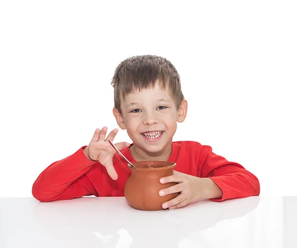 Хворий п'ятирічний хлопчик сидить за білим столом і їсть ложкою з глиняного горщика — стокове фото