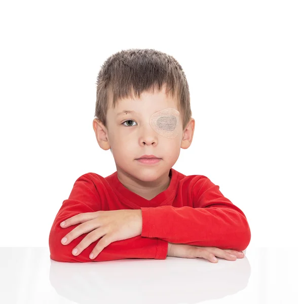 Il bambino di cinque anni siede a un tavolo bianco, l'occhio sinistro è bloccato con un cerotto. — Foto Stock