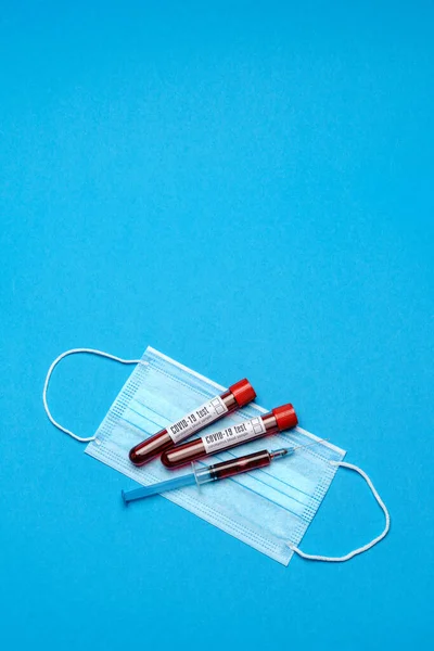 Siringa, provetta con campione di sangue e maschera monouso per prevenire il virus COVID-19 su sfondo blu — Foto Stock