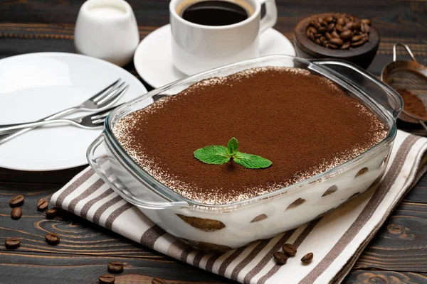 Postre Tiramisú tradicional italiano en bandeja para hornear de vidrio y taza de café expreso caliente fresco sobre fondo de madera — Foto de Stock