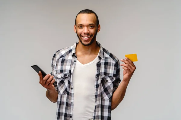 Giovane uomo afro-americano sorridente e zoccolo carta di credito e telefono cellulare su sfondo grigio con spazio di copia — Foto Stock
