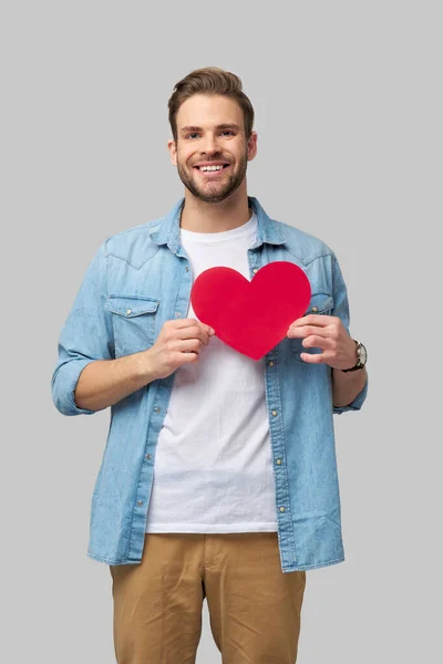 Retrato de hombre encantador de la mano de papel en forma de corazón de la tarjeta de desgaste camisa de estilo casual aislado sobre fondo gris —  Fotos de Stock