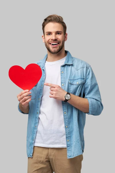 Retrato de hombre encantador de la mano de papel en forma de corazón de la tarjeta de desgaste camisa de estilo casual aislado sobre fondo gris —  Fotos de Stock