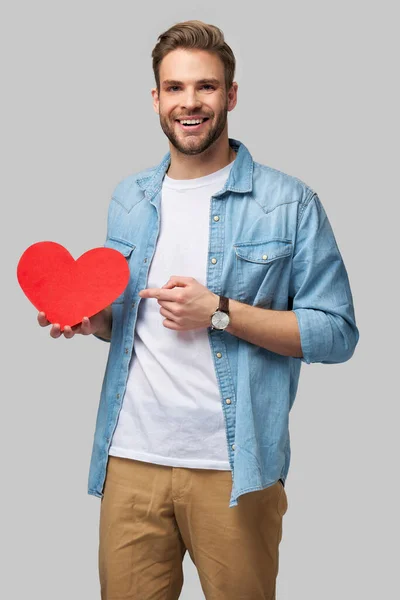 Retrato de hombre encantador de la mano de papel en forma de corazón de la tarjeta de desgaste camisa de estilo casual aislado sobre fondo gris — Foto de Stock