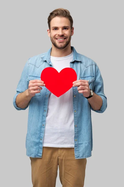 Retrato de hombre encantador de la mano de papel en forma de corazón de la tarjeta de desgaste camisa de estilo casual aislado sobre fondo gris —  Fotos de Stock