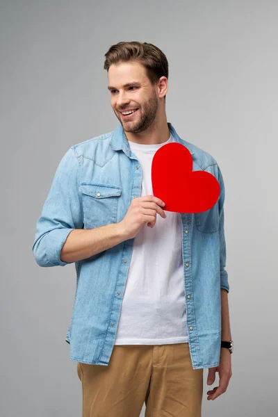 Retrato de homem encantador segurar mão papel coração em forma de cartão desgaste casual estilo camisa isolada sobre fundo cinza — Fotografia de Stock