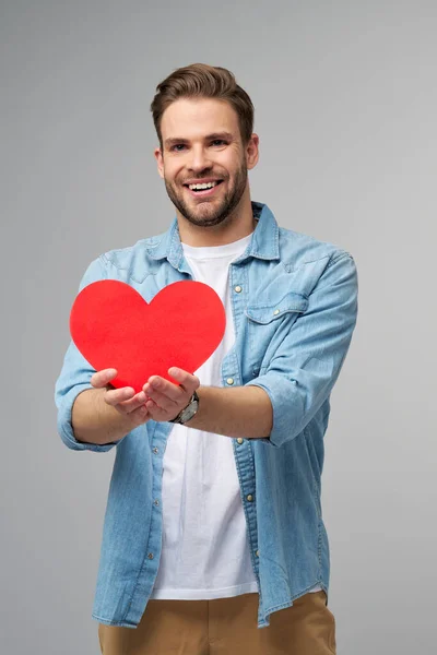 Retrato de hombre encantador de la mano de papel en forma de corazón de la tarjeta de desgaste camisa de estilo casual aislado sobre fondo gris — Foto de Stock