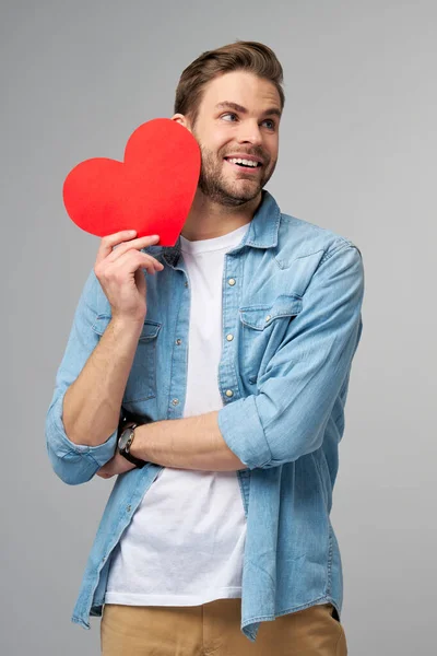 Retrato de hombre encantador de la mano de papel en forma de corazón de la tarjeta de desgaste camisa de estilo casual aislado sobre fondo gris —  Fotos de Stock