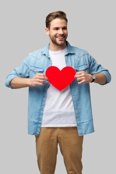 Retrato de hombre encantador de la mano de papel en forma de corazón de la tarjeta de desgaste camisa de estilo casual aislado sobre fondo gris —  Fotos de Stock