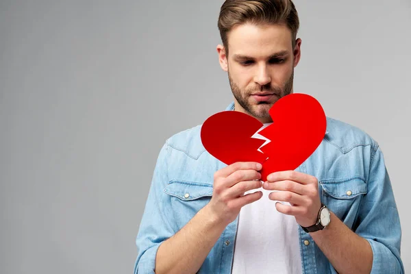 Joven guapo sosteniendo papel roto rojo corazón de San Valentín de pie sobre fondo gris —  Fotos de Stock