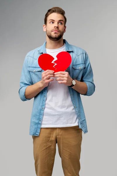 Stilig ung man med brutet papper röd valentine hjärta står över grå bakgrund — Stockfoto