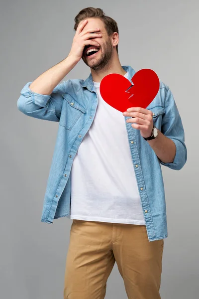 Pohledný mladý muž drží zlomený papír červený valentine srdce stojící nad šedým pozadím — Stock fotografie