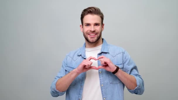 Retrato de hombre caucásico joven guapo haciendo signo de corazón por las manos sobre fondo gris — Vídeos de Stock