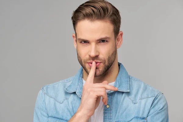 Jovem homem bonito vestindo camisa jeans mantém o dedo nos lábios, pede para não contar informações secretas ou manter o silêncio — Fotografia de Stock