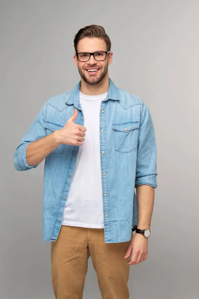 Retrato de jovem bonito caucasiano homem em jeans camisa mostrando grande polegar para cima gesto de pé sobre luz fundo — Fotografia de Stock