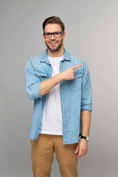 Gelukkig jonge knappe man in jeans shirt wijzend weg staande tegen grijze achtergrond — Stockfoto