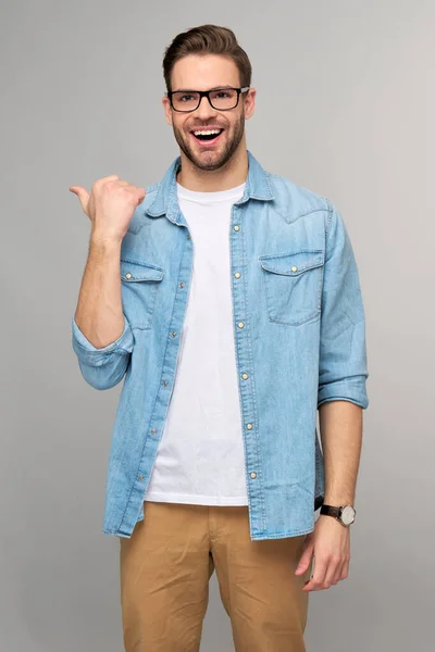 Gelukkig jonge knappe man in jeans shirt wijzend weg staande tegen grijze achtergrond — Stockfoto