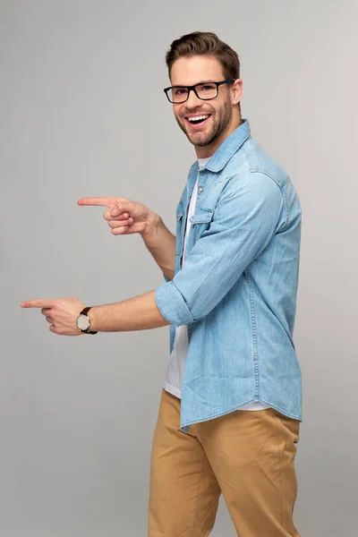 Glücklicher junger gutaussehender Mann in Jeanshemd, der vor grauem Hintergrund wegzeigt — Stockfoto