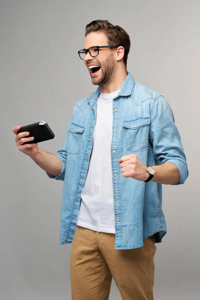 Jovem handcome homem jogando com jogo de vídeo portátil em pé sobre fundo cinza — Fotografia de Stock