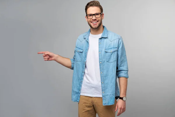 Feliz jovem bonito homem de camisa jeans apontando para longe de pé contra fundo cinza — Fotografia de Stock