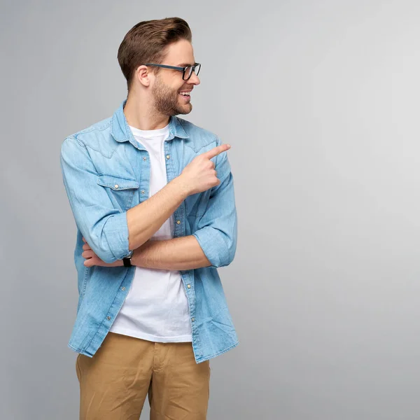 Feliz jovem bonito homem de camisa jeans apontando para longe de pé contra fundo cinza — Fotografia de Stock