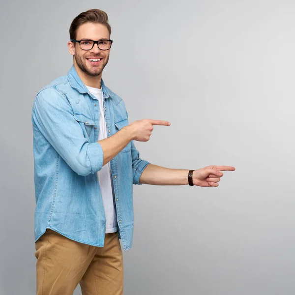 Gelukkig jonge knappe man in jeans shirt wijzend weg staande tegen grijze achtergrond — Stockfoto