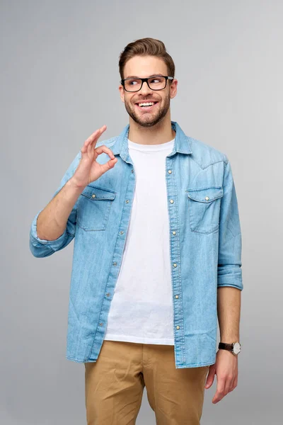 Portret van jonge knappe blanke man in jeans shirt met ok teken gebaar staande over lichte achtergrond — Stockfoto