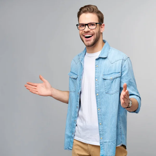 Ung man bär jeans skjorta välkomnar dig med ett leende på ansiktet och hans armar vidöppen stående över grå bakgrund — Stockfoto