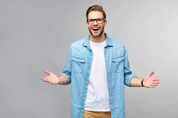 Jeune homme portant une chemise en jean vous accueillant avec un sourire sur le visage et les bras grands ouverts debout sur fond gris — Photo