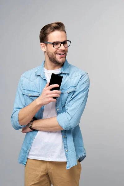 Homem muito casual em camisa jeans azul segurando seu telefone de pé sobre estúdio fundo cinza — Fotografia de Stock