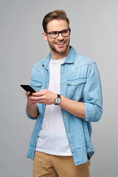 一个穿着蓝色牛仔裤衬衫的非常随意的男人，拿着手机站在工作室灰色的背景上 — 图库照片