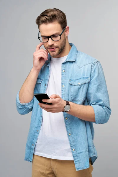 Bastante casual hombre en azul jeans camisa sosteniendo su teléfono de pie sobre estudio gris fondo — Foto de Stock