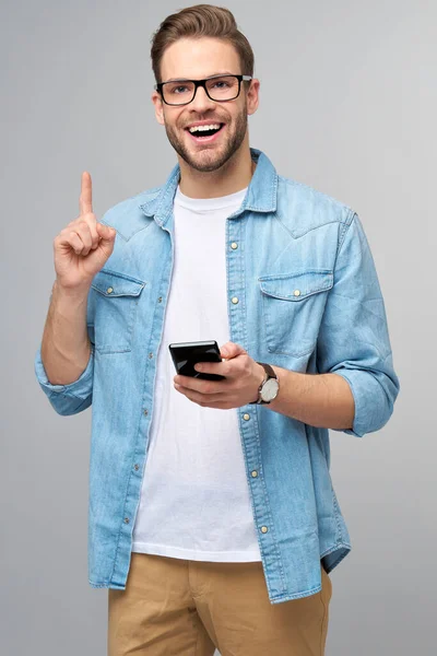 Grazioso casual uomo in blu jeans camicia tenendo il suo telefono in piedi sopra studio grigio sfondo — Foto Stock