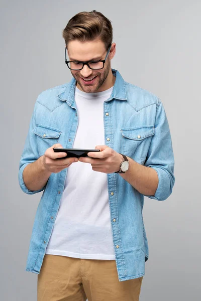 Bastante casual hombre en azul jeans camisa sosteniendo su teléfono de pie sobre estudio gris fondo — Foto de Stock