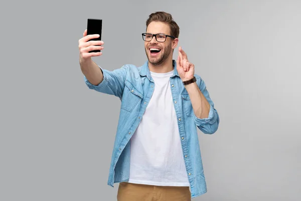 Sorrindo jovem vestindo camisa jeans tirando foto selfie no smartphone ou fazendo chamada de vídeo em pé sobre fundo estúdio cinza — Fotografia de Stock