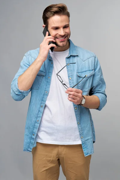 Vrij casual man in blauwe jeans shirt met zijn telefoon staande over studio grijze achtergrond — Stockfoto