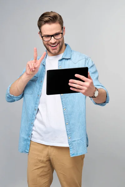 Jeune homme heureux portant une chemise en jean debout et utilisant une tablette sur fond gris studio — Photo