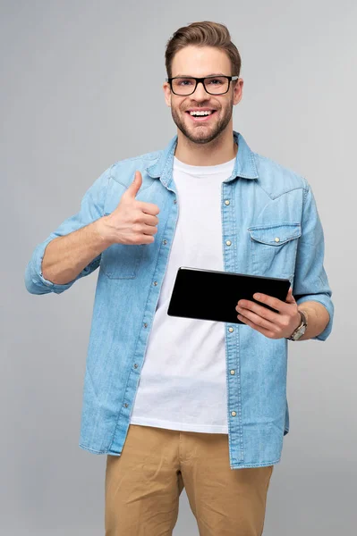 Pria muda yang bahagia dengan kemeja jins berdiri dan menggunakan tablet di atas latar belakang abu-abu studio — Stok Foto