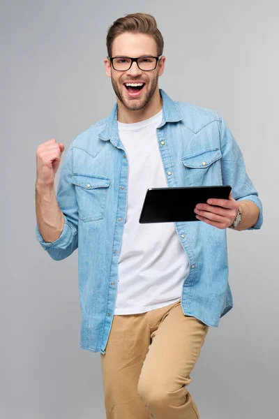 Gelukkig jonge man dragen jeans shirt staande en met behulp van tablet over studio grijze achtergrond — Stockfoto
