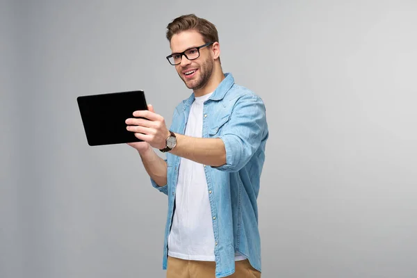 Glücklicher junger Mann in Jeanshemd, stehend und mit Tablet vor studiograuem Hintergrund — Stockfoto