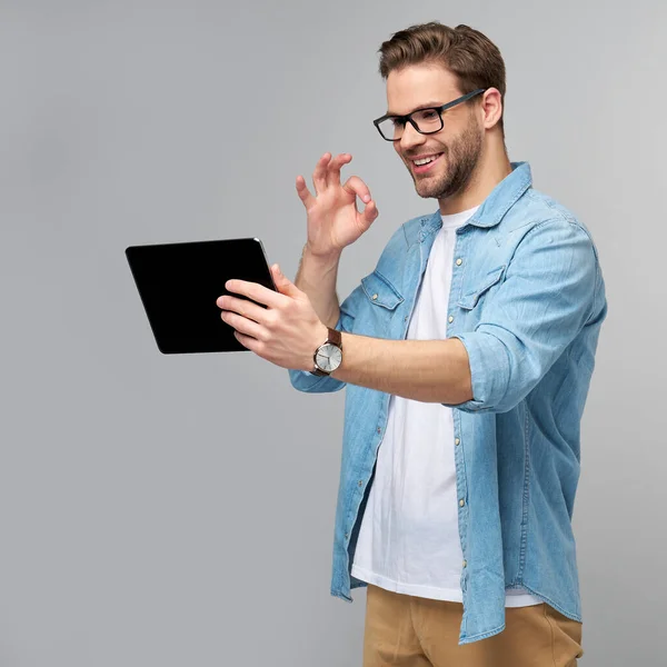 Glücklicher junger Mann in Jeanshemd, stehend und mit Tablet vor studiograuem Hintergrund — Stockfoto