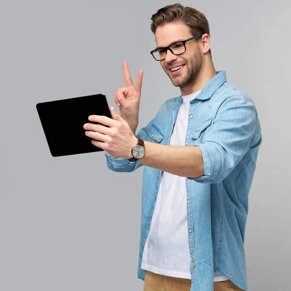 Jovem feliz vestindo camisa jeans de pé e usando tablet sobre estúdio fundo cinza — Fotografia de Stock