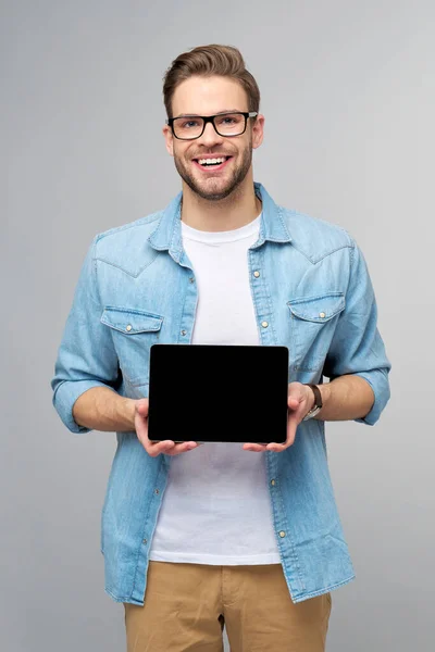 Felice giovane uomo che indossa jeans camicia in piedi e tenendo tablet pc pad su sfondo grigio studio — Foto Stock