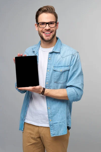 Jovem feliz vestindo camisa jeans de pé e segurando tablet pc pad sobre estúdio fundo cinza — Fotografia de Stock