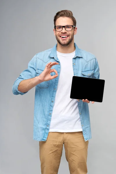 Felice giovane uomo che indossa jeans camicia in piedi e tenendo tablet pc pad su sfondo grigio studio — Foto Stock