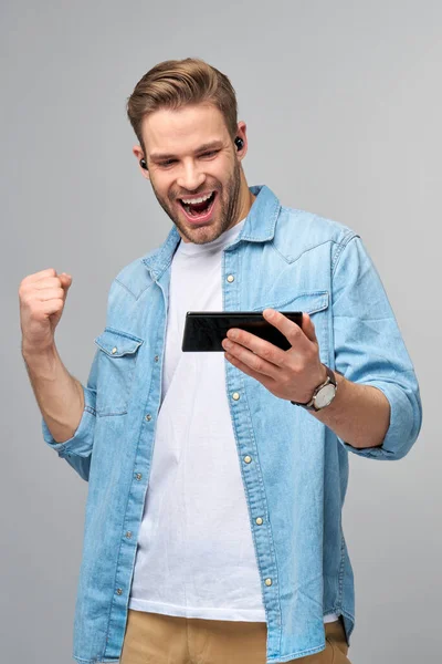Grazioso casual uomo in blu jeans camicia tenendo il suo telefono in piedi sopra studio grigio sfondo — Foto Stock