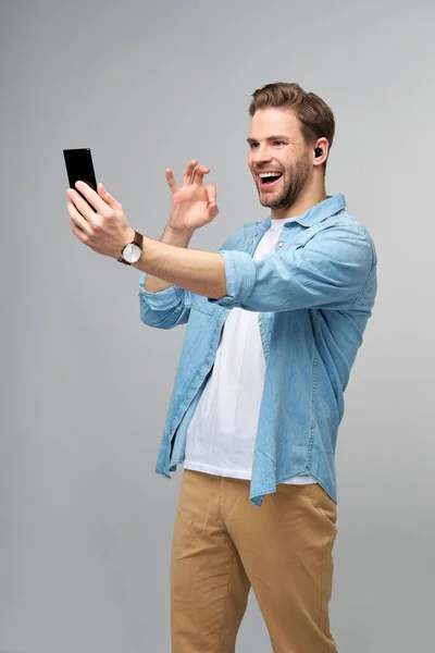 Uśmiechnięty młodzieniec w jeansowej koszuli robiący selfie na smartfonie lub wykonujący wideo połączenie stojące nad szarym tłem studia — Zdjęcie stockowe