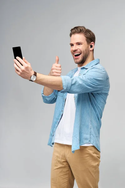 Sorrindo jovem vestindo camisa jeans tirando foto selfie no smartphone ou fazendo chamada de vídeo em pé sobre fundo estúdio cinza — Fotografia de Stock