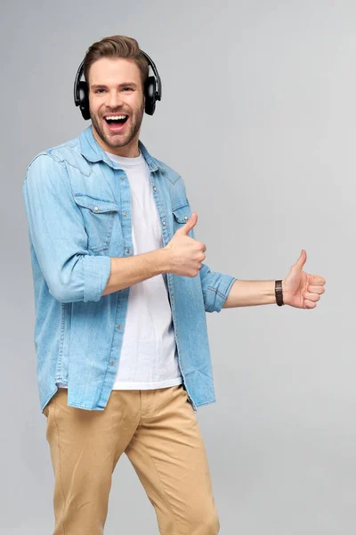 Happy young handsome man in jeans shirt pointing away standing against grey background wearing big headphones — Stock Photo, Image